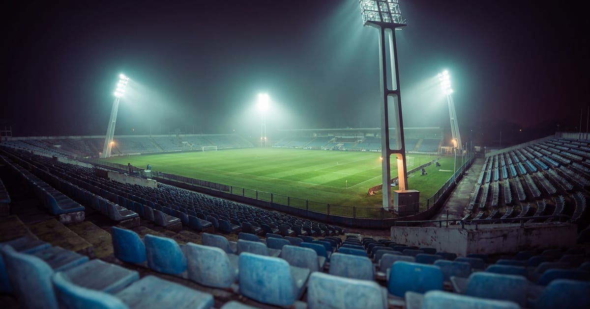 A floodlit stadium at night seen from the empty stands, the pitch bright under the lamps